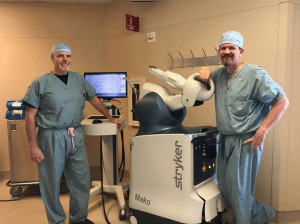 <em>Andrews Sports Medicine &amp; Orthopaedic Center physicians, Drs. Jeffrey Davis and K. David Moore, standing beside the Mako Robotic-Arm Assisted Surgery System used to help patients like missionary Tim Prewitt. “I’d like for people to understand this is not something where I wind up a robot and then go get a cup of coffee,” says K. David Moore, M.D., one of the first surgeons in the area to utilize the innovative technology that allows him to personalize total knee, partial knee and total hip replacement procedures to each of his patients. “Surgeons are still doing the entire operation, it’s just that the robot guides us to make the boning cuts according to our plan for the patient’s individual anatomy.”</em>